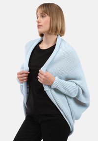 Cardigan lavorato a maglia azzurro chiaro con texture a coste, design aperto sul davanti e vestibilità comoda, indossato sopra una maglietta nera.