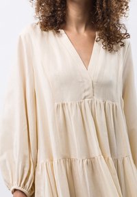 Vestido de capas beige claro hecho de una tela suave. Presenta un escote en forma de V, mangas largas con puños elásticos y un corte relajado. Superficie texturizada.