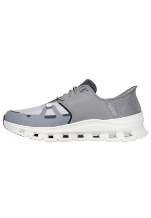 Zapatilla deportiva gris con parte superior de malla, suela blanca acolchada con recortes y un diseño aerodinámico. Acentos en tono gris más oscuro en el talón.
