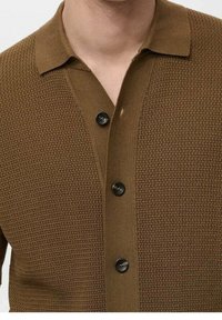 Cárdigan marrón de punto con una superficie texturada, que presenta un cuello con solapa, cinco botones negros y un panel frontal liso en contraste.