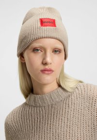Bonnet en tricot beige avec une étiquette rectangulaire rouge portant "HUGO" en texte noir. Le design comprend une texture côtelée et un ajustement serré.