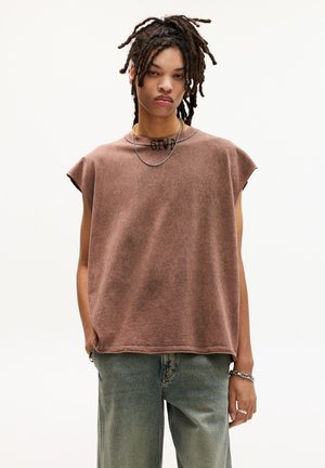 T-shirt marrone oversize senza maniche con scollatura rotonda e bordi grezzi, indossata con jeans blu sbiaditi e arricchita con accessori a catena.