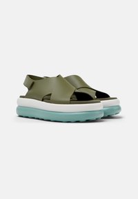 Green sandal with cross straps, light blue sole, and white midsole. Features a back strap for support and an open-toe design.