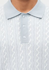 Nahaufnahme eines hellblauen Strick-Poloshirts mit Knöpfen und strukturiertem Zickzackmuster im Stoff, getragen von einer Person.