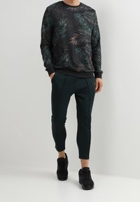 Uomo che indossa una felpa con stampa di foglie tropicali scure, pantaloni neri a 3/4 e sneakers nere, in piedi davanti a uno sfondo chiaro e uniforme.