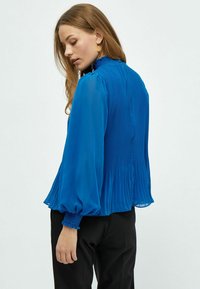 Blusa blu plissettata con maniche lunghe a sbuffo, polsini arricciati e collo alto. Realizzata in tessuto leggero con una texture fluida.