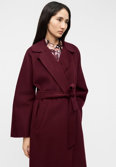 Μπουφάν από μάλλινο ύφασμα σε βουργουνδί απόχρωση με ζώνη στη μέση, κλασικές πέτες και πλαϊνές τσέπες· φορεμένο πάνω από πουκάμισο με λουλουδάτο μοτίβο. Ομαλή υφή, εφαρμοστή γραμμή.