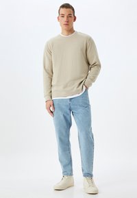 Camicia beige a manica lunga sovrapposta a una maglietta bianca, abbinata a jeans azzurri chiari e sneakers beige. Design semplice con una vestibilità comoda.