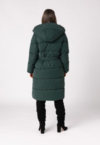 Gevoerde lange jas in donkergroen, met een capuchon, een ceintuur in de taille en een geborduurd ontwerp. Zwarte laarzen tot boven de knie maken de outfit af.