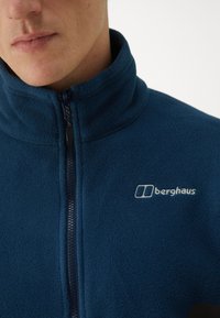 Marineblå fleecejakke med høy krage, hel glidelås og brodert Berghaus-logo på venstre bryst. Myk tekstur, lett design.