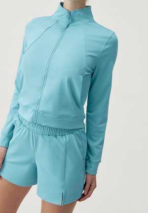 Femme portant une veste zippée bleu clair et un short assorti à taille élastique sur un fond blanc uni.
