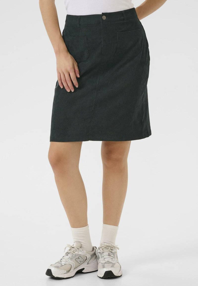 Jupe en denim vert foncé avec une silhouette droite, fermeture à boutons sur le devant et deux poches plaquées. Portée avec des chaussettes blanches et des baskets claires.