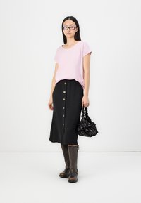 T-shirt rosa a maniche corte abbinato a una gonna midi nera con bottoni dorati, stivali al ginocchio marroni e una borsa a tracolla nera con motivo.
