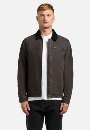 Młody mężczyzna ubrany w ciemnobrązową kurtkę na zamek z czarnym kołnierzem, pod którą ma białą koszulkę i czarne spodnie, stoi na jednolitym tle.