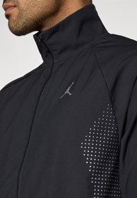 Μαύρο αθλητικό μπουφάν με ψηλό γιακά, κλείσιμο με φερμουάρ, με ασημένιο λογότυπο και διαβάθμιση σε σχέδιο κουκκίδων στη μία πλευρά. Ομαλή υφή.