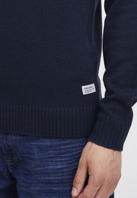 Pulover tricotat bleu marin, cu tiv și manșete cu ață. Are un material texturat și un mic etichetă de marcă în partea stângă jos.