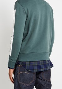 Grön sweatshirt med vit randdetalj längs vänster arm, ribbade ärmslut och nederkant, lagd över en rutig skjorta i blått och grönt.