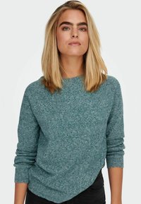 Maglione in maglia teal con maniche lunghe e scollatura arrotondata. Texture morbida con un sottile motivo melangiato. Abbinato a pantaloni neri.