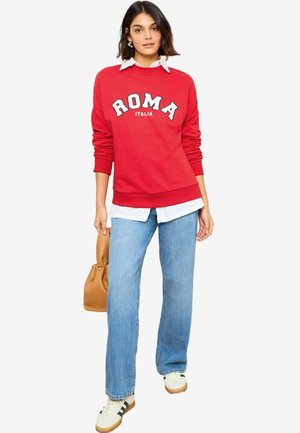 Rode sweater met de tekst "ROMA", witte blouse eronder, blauwe jeans en een tan handtas. Casual sneakers met groene strepen. Zacht van textuur.
