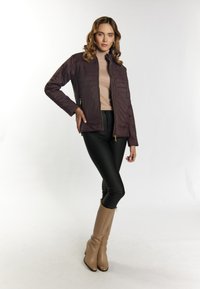 Giacca imbottita di un viola scuro con zip frontale, colletto alto e tasche laterali. Indossata sopra un top beige e pantaloni neri, abbinata a stivali beige.