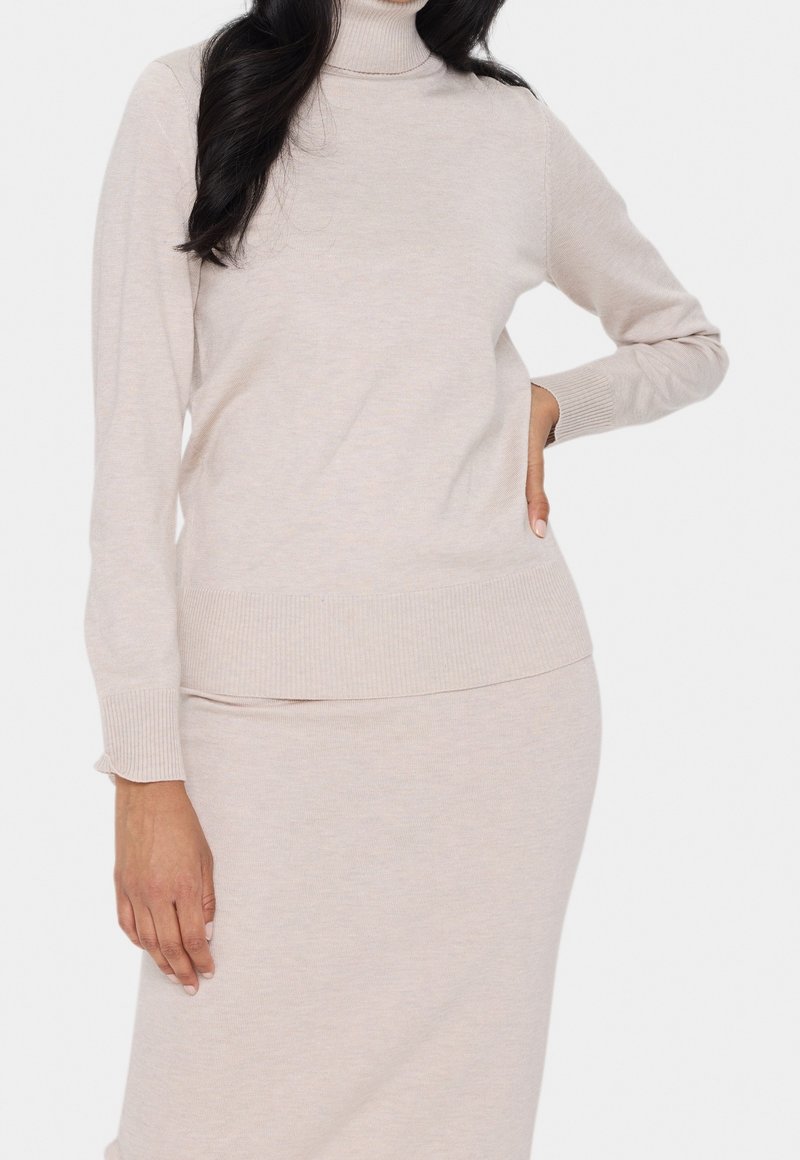 Femme portant un pull à col roulé beige clair et une jupe ajustée assortie, posant avec une main sur la hanche devant un fond uni.
