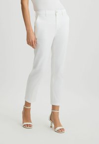 Pantalones blancos a medida con un ajuste slim, cuentan con un frente plano, cierre de un solo botón y bolsillos laterales. Combinados con sandalias blancas de tacón alto.