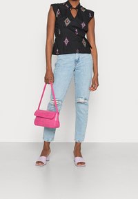 Donna con top senza maniche nero con motivi colorati, jeans strappati chiari, sandali con tacco lilla, mentre regge una borsa di colore rosa acceso.
