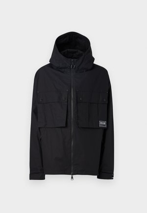 Veste imperméable noire avec capuche, fermeture éclair à l'avant, deux grandes poches poitrine et logo. Elle possède une coupe décontractée et des poignets réglables.