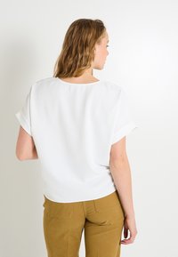 Blusa blanca de manga corta con cuello redondo, corte relajado y mangas enrolladas. Textura suave, combinada con pantalones marrones.