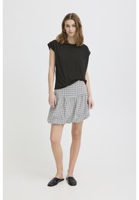 Ženska stoji, oblečena v črno majico s kratkimi rokavi, belo in črno karo krilo, črne natikače in srebrne obroče za ušesa.