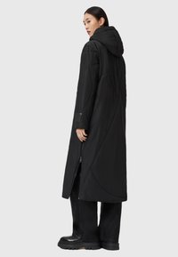 Długi czarny puchowy płaszcz z kapturem, z geometrycznym przeszyciem, z zamkiem błyskawicznym z przodu i bocznymi kieszeniami. Nosi się go na czarne spodnie o szerokich nogawkach i buty.