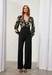Blusa preta com padrão floral e detalhes em dourado, combinada com calças pretas de cintura alta, com pernas largas e sandálias pretas de ponta aberta.