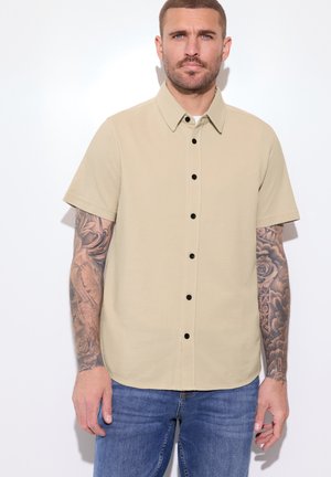 Man met kort haar en tatoeages op beide armen, gekleed in een beige kort mouwen overhemd met knopen en blauwe spijkerbroek tegen een witte achtergrond.