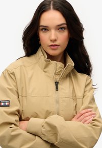 Veste zippée beige avec un col montant et des poignets élastiques, présentant un logo sur la manche gauche et un tissu lisse et texturé.