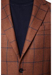Brązowa marynarka w wzory z granatowym golfem pod spodem. Marynarka ma teksturowany materiał, ostre klapy i zapięcie na jeden guzik.