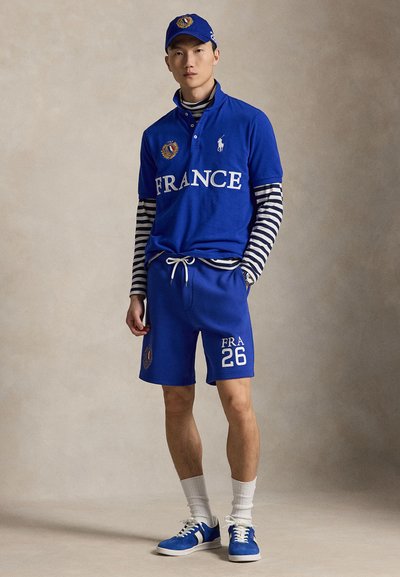 Joven vestido con ropa deportiva azul de Francia, incluyendo gorra, camiseta polo, pantalones cortos, camiseta interior de manga larga a rayas, calcetines blancos y zapatillas deportivas azules.