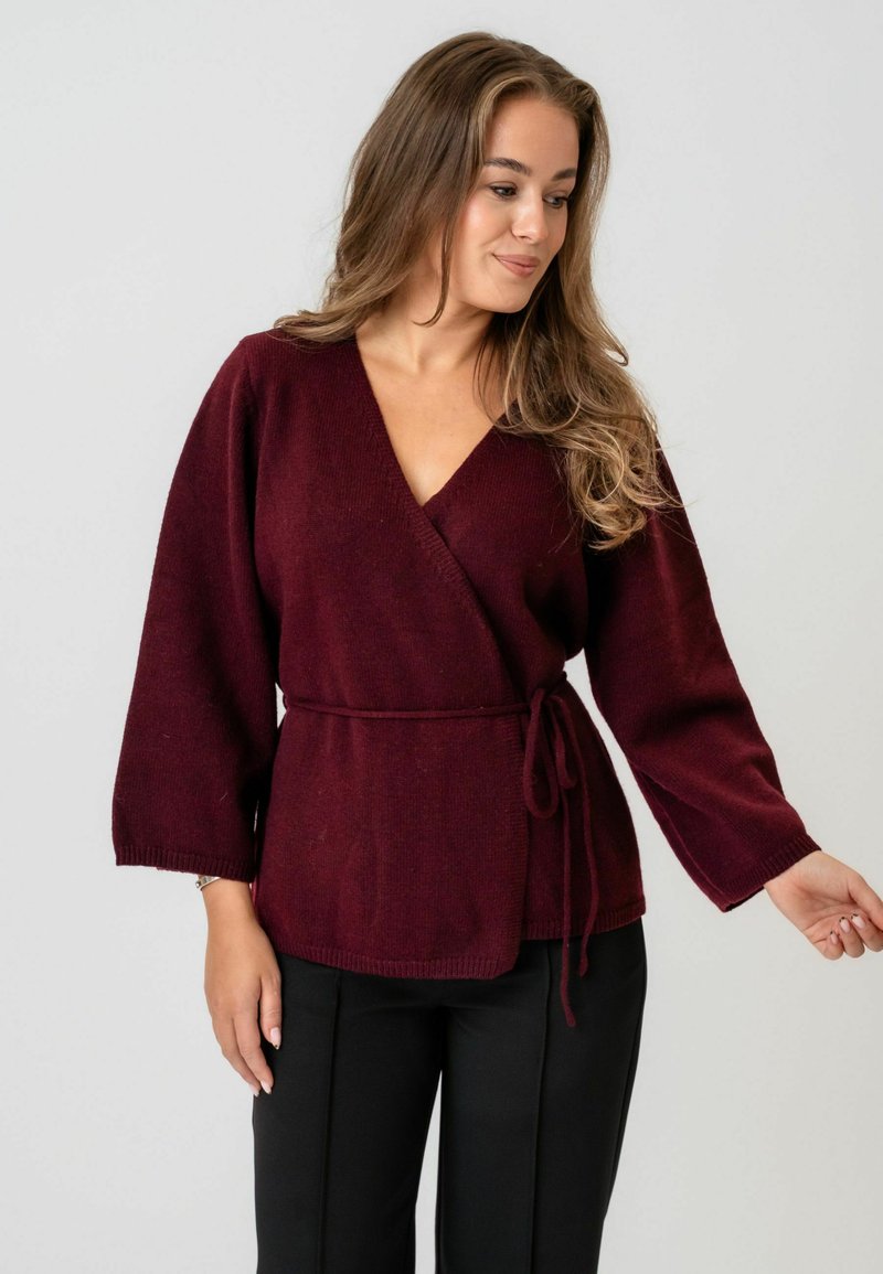 Pullover bordeaux cu decolteu adânc în V, mâneci largi și talie legată. Textură moale și manșete cu model cu nervuri care întregesc designul. Asortat cu pantaloni negri.