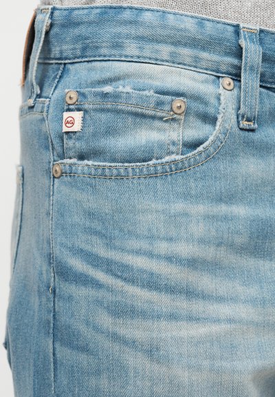 Τσέπη από ελαφρύ γαλάζιο τζιν με μικρή υφασμάτινη ετικέτα που εμφανίζει το λογότυπο "AG" και ορατές ραφές και καρφιά.