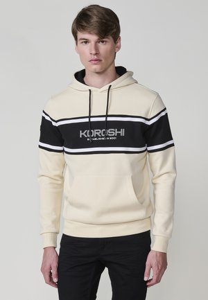 Hoodie u krem boji s crnom prugom preko prsa, s nazivom brenda u srebrnoj boji i prednjim džepom.