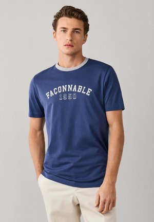 Jeune homme aux cheveux courts portant un T-shirt bleu avec col blanc et texte blanc "FAÇONNABLE 1950", debout avec une main dans la poche.