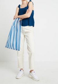 Námořnický žebrovaný top s mašlovými ramínky, bílé zkrácené džíny a bílé tenisky; drží průhlednou modro-pruhovanou tašku.