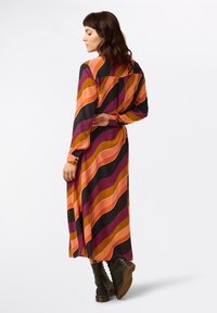 Langes, gemustertes Kleid mit wellenförmigen Streifen in Orange, Lila und Schwarz, mit geknöpften Manschetten und einem lässigen Schnitt, kombiniert mit schwarzen Stiefeln.