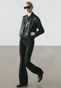 Woman wearing black leather jacket, white turtleneck, black flared pants, black shoes, large black sunglasses, walking with hands in pockets.