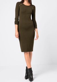 Olivgrön, figursydd stickad klänning med långa ärmar, ribbad textur och dekorativa detaljer på axlarna. Bärs med svarta ankelskor.