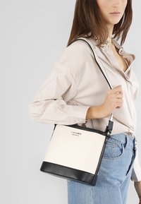 Vit och svart strukturerad handväska med en texturerad svart bas, justerbar rem och tryckt logotyp på framsidan.