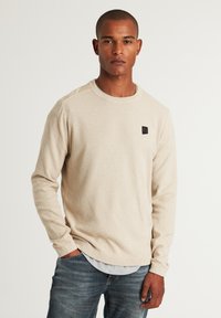 Beige strukturierter Pullover mit langen Ärmeln, rundem Ausschnitt und einem kleinen schwarzen Logo-Patch auf der Brust. Trägt man über einem hellen Hemd mit Kragen.