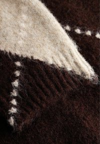 Tamno smeđa i kremasta pletena vuna s teksturiranim uzorkom. Sadrži geometrijski dizajn s uzdignutim šavovima i mekanim, plišanom završnom obradom.
