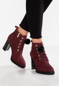 Botas de tobillo de ante burdeos con cordones y cierre de cremallera, acentuadas por pompones negros y un tacón cuadrado brillante, con una suela texturizada.