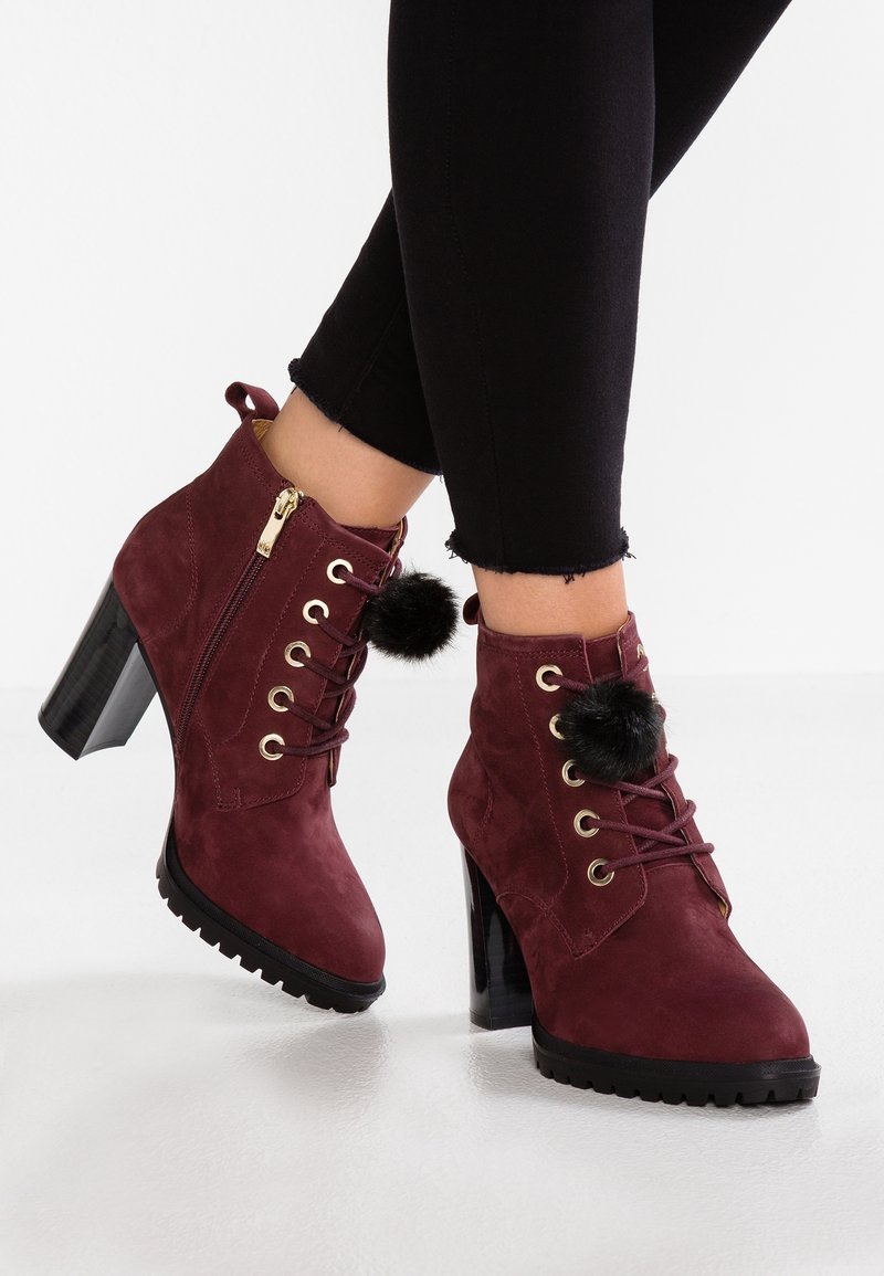 Botas de tobillo de ante burdeos con cordones y cierre de cremallera, acentuadas por pompones negros y un tacón cuadrado brillante, con una suela texturizada.