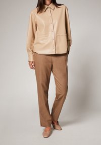 Chemise boutonnée en simili cuir beige avec un col, associée à un pantalon marron ajusté et des chaussures beiges à bout fermé, avec des fentes latérales discrètes.